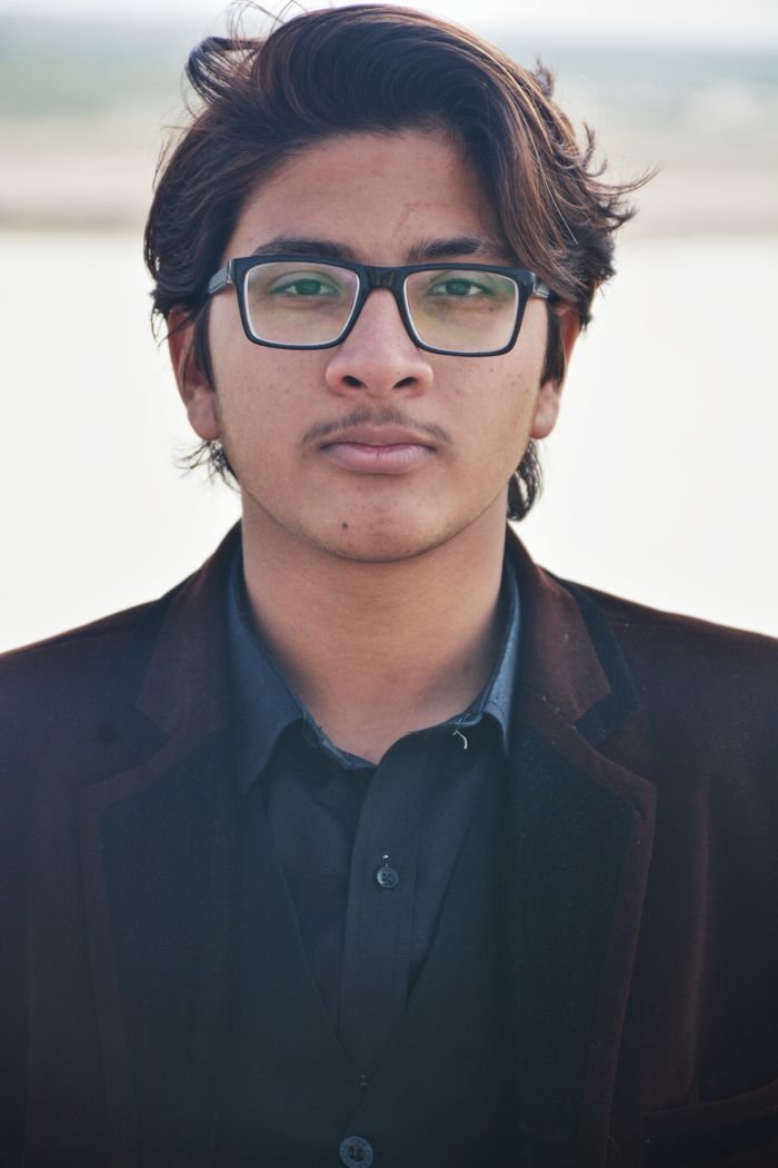 A young man with eyeglasses and a blazer, gazing confidently. Outdoors setting.